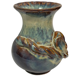 May include: A ceramic vase with a flared rim and a rounded body, featuring a glossy glaze in shades of deep brown, sage green, and teal. A sculpted, rope-like decorative element wraps around the midsection, ending in a shell-like motif.