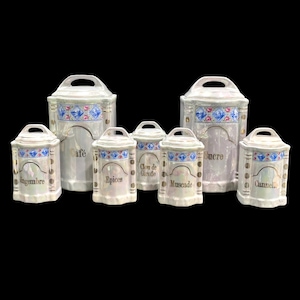 May include: A set of seven iridescent white ceramic canisters with arched handles and blue and pink floral bands. The canisters are labeled in gold text: Gingembre, Café, Epices, Clou de Girofle, Muscade, Sucre, and Cannelle, featuring vertical gold accents.