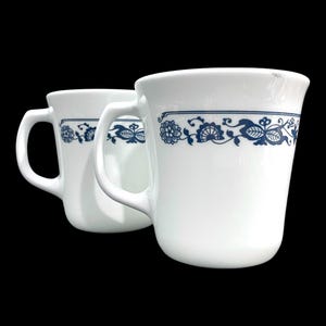 Può includere: Due tazze in ceramica bianca con un motivo floreale blu. Le tazze hanno un manico curvo e un bordo largo. Il motivo floreale blu è un motivo ripetuto attorno alla parte superiore della tazza. Le tazze sono su uno sfondo nero.