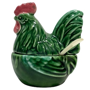 Może przedstawiać: Ceramiczny pojemnik w kształcie koguta z pokrywką i małą łyżeczką. Pojemnik jest błyszczący, głęboko zielony z czerwonym grzebieniem i koralami. Łyżeczka jest w kolorze ecru. Projekt jest szczegółowy i teksturowany.