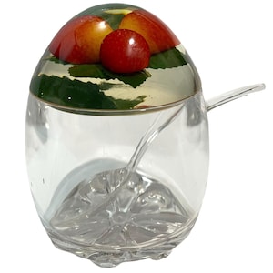 Può includere: Una zuccheriera in vetro trasparente con un coperchio decorativo con frutta rossa e gialla su sfondo verde. Un cucchiaio trasparente è all'interno della ciotola. La ciotola ha una forma unica e arrotondata.