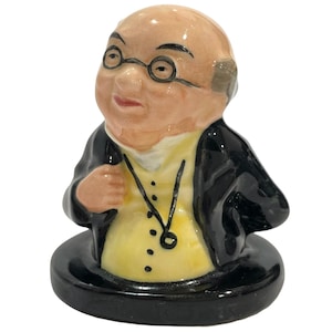 Peut inclure: Une figurine en céramique représentant une personne âgée chauve portant des lunettes rondes, un gilet jaune avec une chaîne noire et une veste noire. Le personnage est présenté en buste sur une base circulaire noire à la finition brillante.