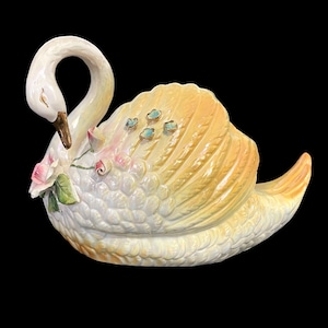 May include: A white porcelain swan figurine with a yellow and pink glaze. The swan has a detailed design with feathers and a floral pattern. The swan's head is tilted down and its beak is open. The swan is sitting on a base with a floral design.