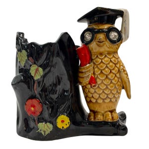 Puede incluir: Una figura de búho de cerámica con gorro de graduación y gafas, sosteniendo un pergamino rojo. El búho es de color canela con detalles marrones y está junto a un tocón de árbol negro con detalles florales coloridos. Una pieza decorativa única.