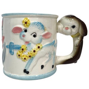 May include: A white ceramic mug featuring a raised relief of a blue lamb wearing a yellow flower garland. The mug has a light blue rim and base, and the handle is shaped like a brown and white puppy dog head with a yellow collar.