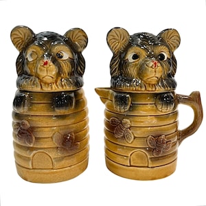 Può includere: Un set di vasetto di miele e panna in ceramica. Il vasetto di miele e la panna sono a forma di alveare con un coperchio a testa di orso. Il vasetto di miele ha un coperchio a testa di orso e la panna ha un manico e un beccuccio. Il set è marrone e beige con dettagli di api e foglie.