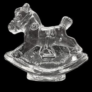 May include: A clear glass rocking horse figurine. The horse has a detailed mane, bridle, and saddle. The base is curved, and the entire piece is transparent, with a glossy finish. The background is black.
