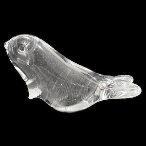 Può includere: Una scultura in vetro trasparente di una foca, sdraiata con le pinne estese. La foca ha un corpo liscio e arrotondato e un viso dettagliato. La scultura è ambientata su uno sfondo nero.