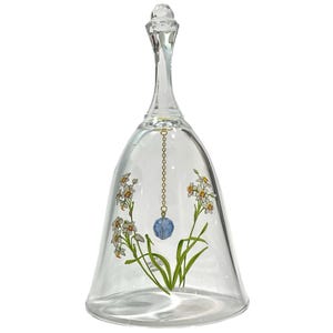 Op de afbeelding: Een heldere glazen bel met een geschilderd bloemmotief van witte en gele narcissen met groene stelen. De bel heeft een hoog, slank handvat en een goudkleurige ketting die aan de binnenkant hangt en eindigt in een gefacetteerde blauwe kraal.