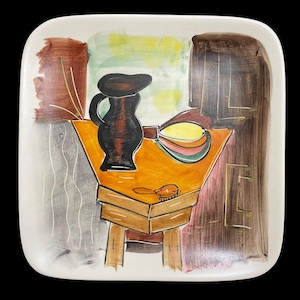 Puede incluir: Un plato cuadrado de cerámica con una escena de bodegón pintada a mano. El plato presenta una jarra marrón, una fruta amarilla y verde y una mesa de madera con un cepillo en la parte superior. El fondo es un diseño abstracto colorido.