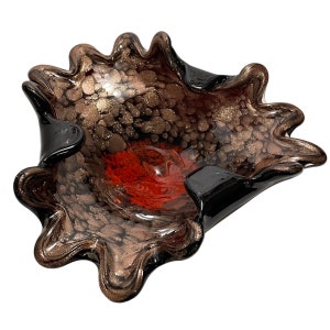 Puede incluir: Un cuenco decorativo de cristal con un borde ondulado irregular. El cuenco presenta un centro rojo intenso que se transforma en un patrón moteado de color marrón oscuro y negro con motas doradas metálicas por todo el cristal, creando un aspecto texturizado.