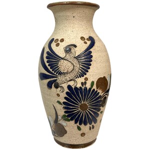 May include: A beige ceramic vase with a speckled finish, featuring a hand-painted design of a blue bird with wings spread and a large dark blue flower. The vase has a brown rim and base, with additional small brown and green floral accents.