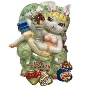 May include: A ceramic figurine of a white rabbit character with blonde hair sitting in a green armchair, holding a chocolate sundae. The chair features the text I LOVE CHOCOLATE FUND, with a box of chocolates and a bag labeled CHOCOLATE SMOOCHS.