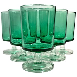 May include: A set of six matching emerald green glass goblets, each featuring a cylindrical bowl, a short stem, and a circular base. The glassware is arranged in a triangular formation, showcasing the translucent green color and smooth finish.