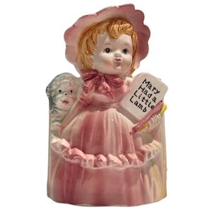 May include: A ceramic figurine of a child wearing a pink bonnet and dress, holding a white rectangular sign with the black text Mary Had a Little Lamb. A small white lamb figure stands beside the child, who has blonde hair and a pink bow.