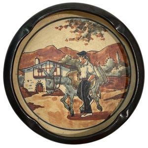 May include: Round ceramic ashtray with a black rim and a painted scene. The scene depicts a man leading a donkey carrying supplies near a house and mountains. The color palette includes brown, beige, and black.
