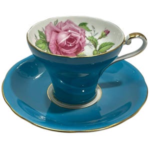 Puede incluir: Un juego de taza de té y platillo de porcelana color verde azulado con borde y asa dorados. El interior de la taza muestra una ilustración detallada de una gran rosa rosa con hojas verdes y un pequeño capullo sobre un fondo blanco.