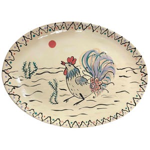 May include: An oval ceramic platter with a cream-coloured base featuring a hand-painted rooster in the centre. The rooster has a red comb, blue and white feathers, and a colourful tail. The border has a black zigzag pattern with small dots.