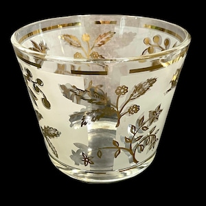 Könnte beinhalten: Ein klares Glas mit goldenen Blumen- und Blattmustern. Das Glas hat einen mattweißen Hintergrund mit goldenen Akzenten. Der Rand und der Boden des Glases sind ebenfalls mit Gold verziert. Das Glas steht vor einem schwarzen Hintergrund.