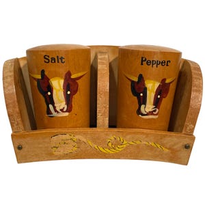 May include: A wooden salt and pepper shaker set housed in a matching curved wooden caddy. Each cylindrical shaker features a painted cow head design and the words Salt or Pepper in black lettering. The caddy includes a yellow rope graphic.