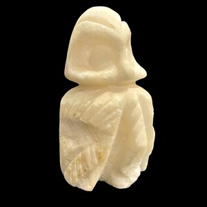 May include: A carved alabaster sculpture of a stylized figure with a draped garment and a head covering. The off-white sculpture has detailed carving and is set against a black background.