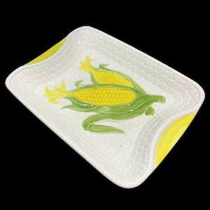 Könnte beinhalten: Weiße Keramik-Servierplatte mit gewebtem Muster und einem Reliefbild von zwei Maiskolben mit gelben Körnern und grünen Hüllen.