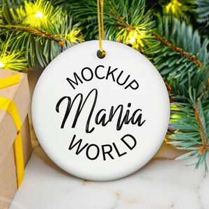 May include: White ceramic Christmas ornament with black text that reads "Mockup Mania World" hanging from a gold string. The ornament is on a white marble surface with a brown gift box wrapped in yellow ribbon.