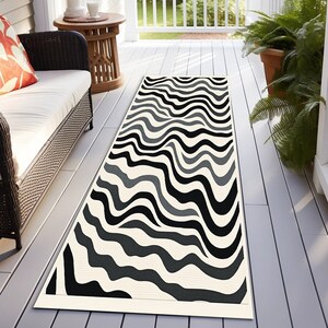May include: A rectangular outdoor rug with a wavy pattern in black, gray, and off-white. The rug is placed on a wooden deck, and the design creates a visual flow. The rug's dimensions are suitable for a porch or patio.
