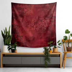 May include: A red tapestry with a distressed, textured design hangs on a white wall above a gray media console with wooden legs. The console is decorated with potted plants, including a snake plant, a monstera, and a fern.