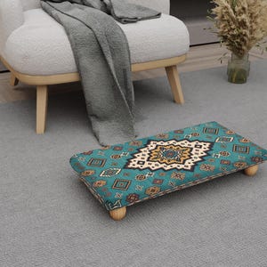 May include: A rectangular footstool with a teal and patterned design. The pattern includes geometric shapes in shades of brown, gold, and white. The footstool has four short, rounded wooden legs. The footstool is placed on a grey rug.