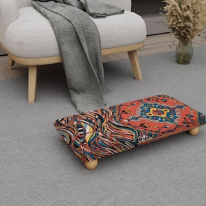 May include: A rectangular footstool with a colorful patterned design. The footstool has a wooden frame and four short, rounded legs. The top features a mix of red, blue, and yellow patterns. The footstool is placed on a gray carpet.