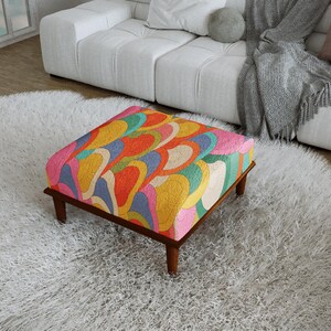 May include: A square ottoman featuring a vibrant, multi-colored pattern of overlapping, curved shapes in shades of orange, yellow, blue, green, and pink. The ottoman is supported by four dark brown wooden legs and has a textured, quilted surface.