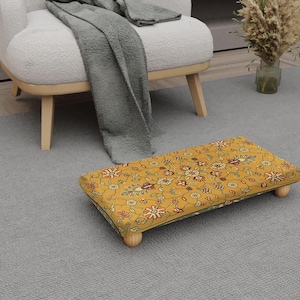 May include: A rectangular footstool with a yellow floral pattern, resting on four small wooden legs. The footstool is placed on a gray carpet, near a white armchair and a gray blanket.