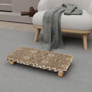 May include: A rectangular footstool with a patterned rug design. The rug features a beige background with floral and geometric patterns in shades of brown, cream, and red. The footstool is supported by four small, rounded wooden legs.
