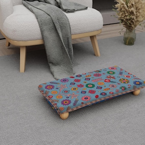 May include: A rectangular footstool with a blue floral pattern, featuring various colorful flowers and geometric shapes. The stool has four wooden legs and a decorative border. The footstool is placed on a gray carpet near a white chair.