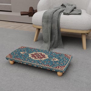 May include: A rectangular footstool with a teal and red floral pattern. The footstool has a wooden frame and four rounded wooden legs. The design features a central medallion and floral motifs. The footstool is placed on a grey carpet.