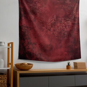 May include: A red tapestry with a faded floral pattern hangs on a white wall above a wooden shelf and a dark gray media console with three drawers. A small potted plant and a stack of books are on the console.