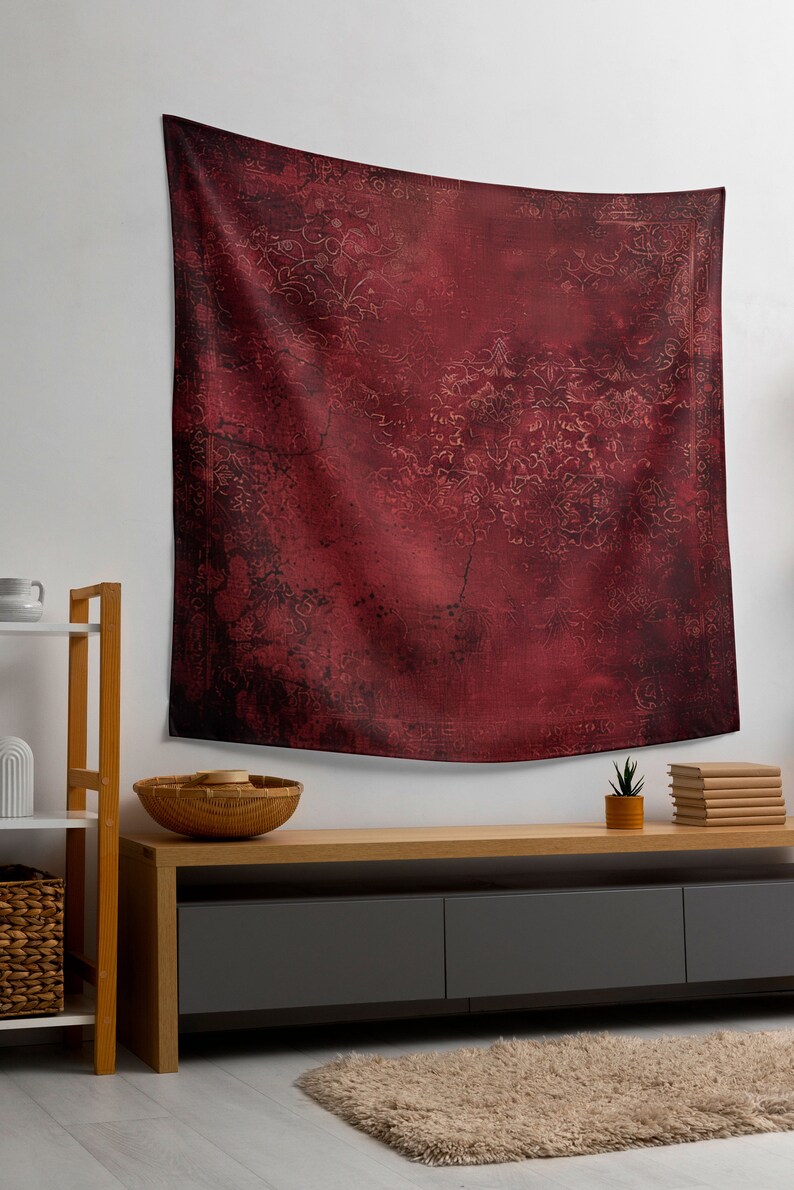 May include: A red tapestry with a faded floral pattern hangs on a white wall above a wooden shelf and a dark gray media console with three drawers. A small potted plant and a stack of books are on the console.