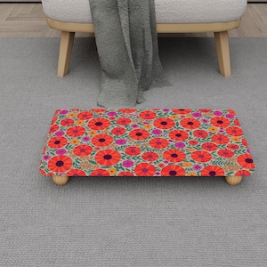 May include: A rectangular footstool with a floral pattern featuring red, orange, and pink flowers on a cream background. The footstool has four wooden legs and is placed on a gray carpet.