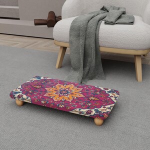 May include: A rectangular footstool with a vibrant floral pattern in shades of pink, orange, and blue. The footstool is supported by four small, rounded wooden legs. The design is reminiscent of a traditional rug.