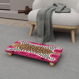 May include: A pink footstool with a leopard print design. The footstool has a rectangular shape with rounded corners and is supported by four wooden legs. The leopard print features brown spots on a cream background, with pink and black accents.