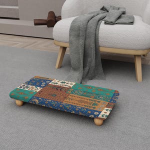 May include: A rectangular footstool with a patchwork design in shades of blue, teal, brown, and cream. The footstool has rounded corners and is supported by four short, light-colored wooden legs. The design features floral and geometric patterns.