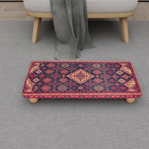 May include: A rectangular footstool with a patterned rug design. The rug features a navy blue background with red and peach-coloured geometric patterns. The footstool has four rounded wooden legs.