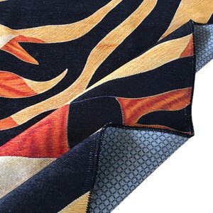 a close-up view of a vibrant, abstract rug with a striking pattern of black, orange, and yellow colors. The rug appears to be made of a soft, plush material, and it is folded or rolled up, revealing its intricate design.