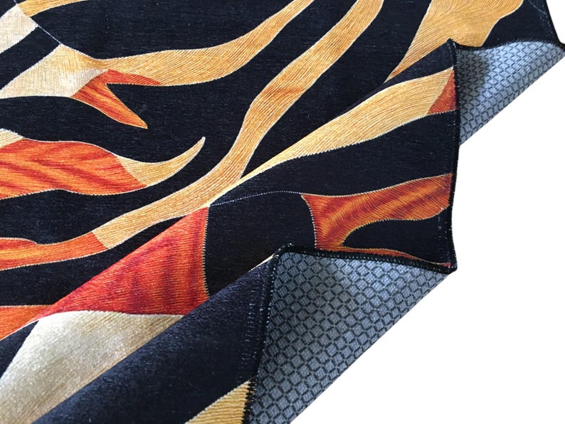 a close-up view of a vibrant, abstract rug with a striking pattern of black, orange, and yellow colors. The rug appears to be made of a soft, plush material, and it is folded or rolled up, revealing its intricate design.