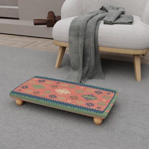 May include: A rectangular footstool with a patterned top in shades of red, green, and blue, supported by four rounded wooden legs. The footstool is placed on a gray carpet, near a white armchair.