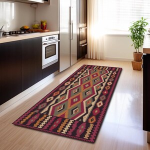 Peut inclure: Un tapis de cuisine rectangulaire avec un motif géométrique dans les tons bordeaux, vert, beige et orange. Le tapis présente un motif de losanges et de zigzags, ajoutant une touche de couleur à l'espace cuisine. Le tapis est posé sur un sol en bois clair.