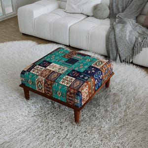 Puede incluir: Un otomano cuadrado con una parte superior de tela de retazos multicolor con intrincados patrones geométricos y florales en tonos azul verdoso, azul marino, naranja y crema, apoyado sobre cuatro patas cortas de madera marrón oscuro con acabado liso.