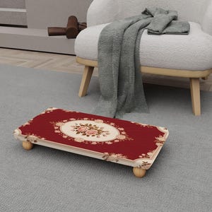May include: A rectangular footstool with a red surface and a floral design. The footstool has a cream-colored border and is supported by four small, rounded wooden legs. The center of the footstool features an oval floral pattern.