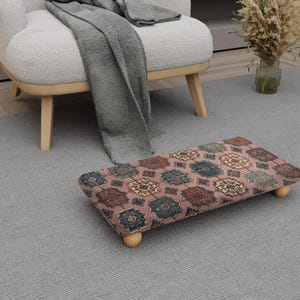 May include: A rectangular footstool with a patterned top in shades of pink, brown, and teal, resting on four short, light-colored wooden legs. The footstool is placed on a gray rug near a white armchair and a gray blanket.